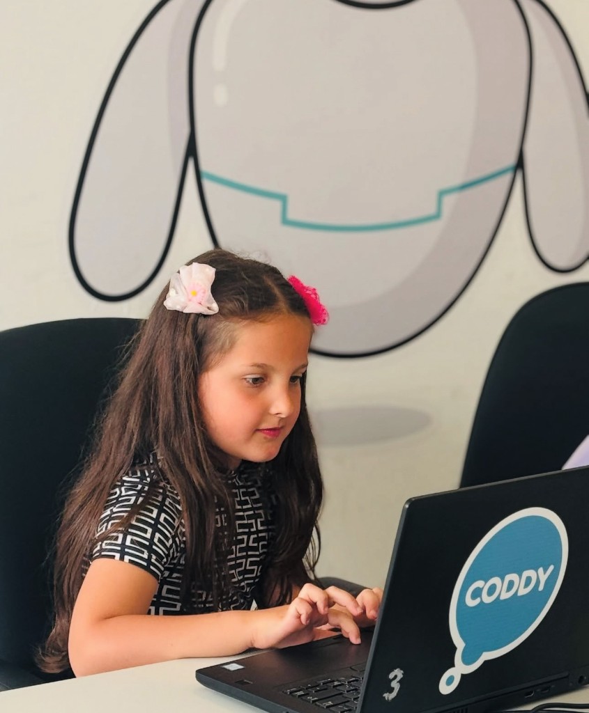 Vajzë e fokusuar në laptop me ngjitëse Coddy, në klasë me mural roboti